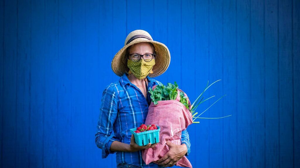 Lisa Miskelly wears a mask and a sun hat while holding produce, against a bright blue background