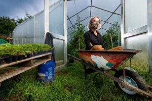 Lisa Miskelly sits wearing a mask in a wheelbarrow with greenhouse in background