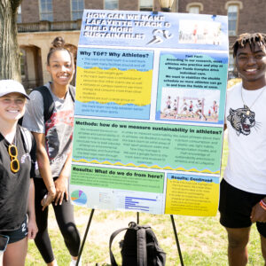 Three students smile at the camera in front of a poster titled "How can we make Lafayette track and field more sustainable?"