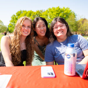 Three students smile at the camera across a table with a red tablecloth