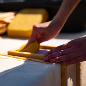 Two hands reach down to smooth a sponge across a recycled paper mixture in a wooden frame