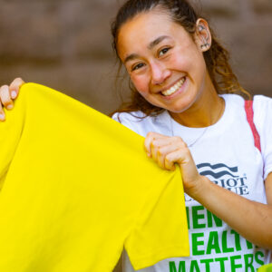 A student smiles at the camera and holds up a yellow t-shirt
