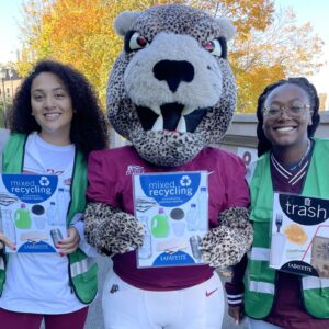 Two students in green vests stand on either side of the Leopard mascot, all holding trash and recycling signage