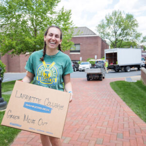 A student in a green t-shirt smiles at the camera and walks down a red brick path while holding a box that says "Lafayette College Green Move Out"