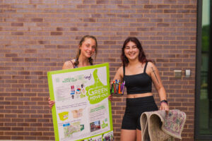 Two students smile at the camera. The student on the left holds up a Green Move Out information sign while the student on the right holds a container of writing utensils and a rug.