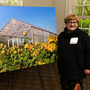 A woman smiles at the camera while standing in front of a poster of yellow flowers surrounding LaFarm's Greenhouse