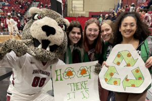 Four students in green vests smile at the camera next to the Leopard mascot while holding recycling signage.