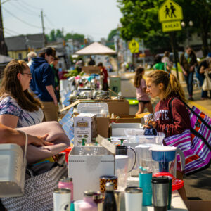 Students and community members stand on either side of a long row of tables covered in boxes and items for sale.