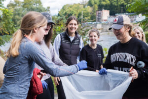 A group of smiling students with the river behind them hold open a clear trash bag.