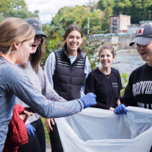 A group of smiling students with the river behind them hold open a clear trash bag.