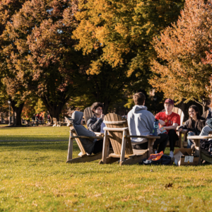 A group of students sit in a circle on Adirondack chairs with a fall backdrop
