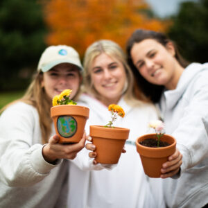 Three students smile at the camera and hold out flowers in brown pots